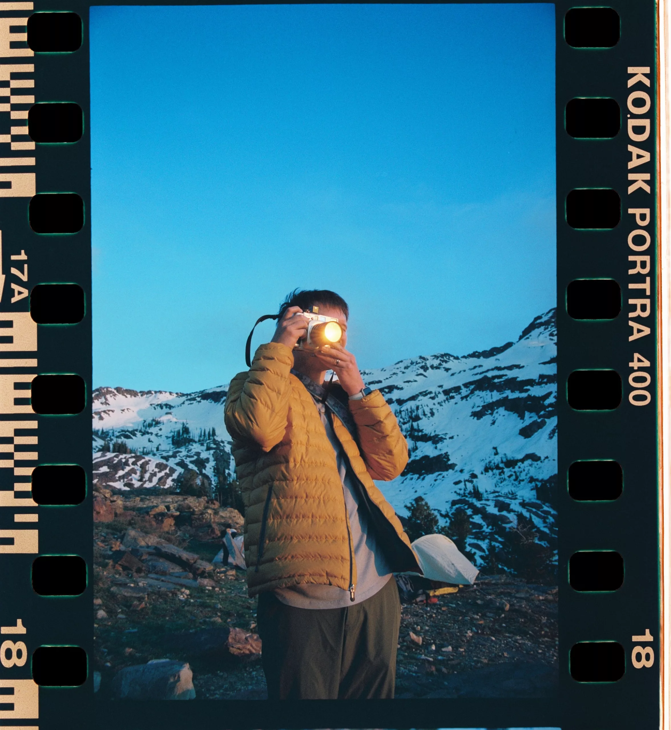 Full 35mm Portra 400 frame with sprockets: a man raising a camera before snowy mountains
